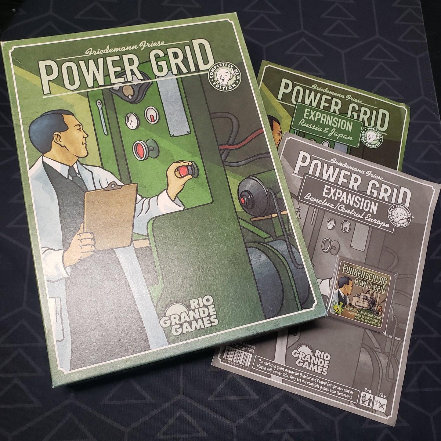 Image shows the front cover of the box of the Power Grid board game with the instructions for two board expansions and some promo cards sitting next to it