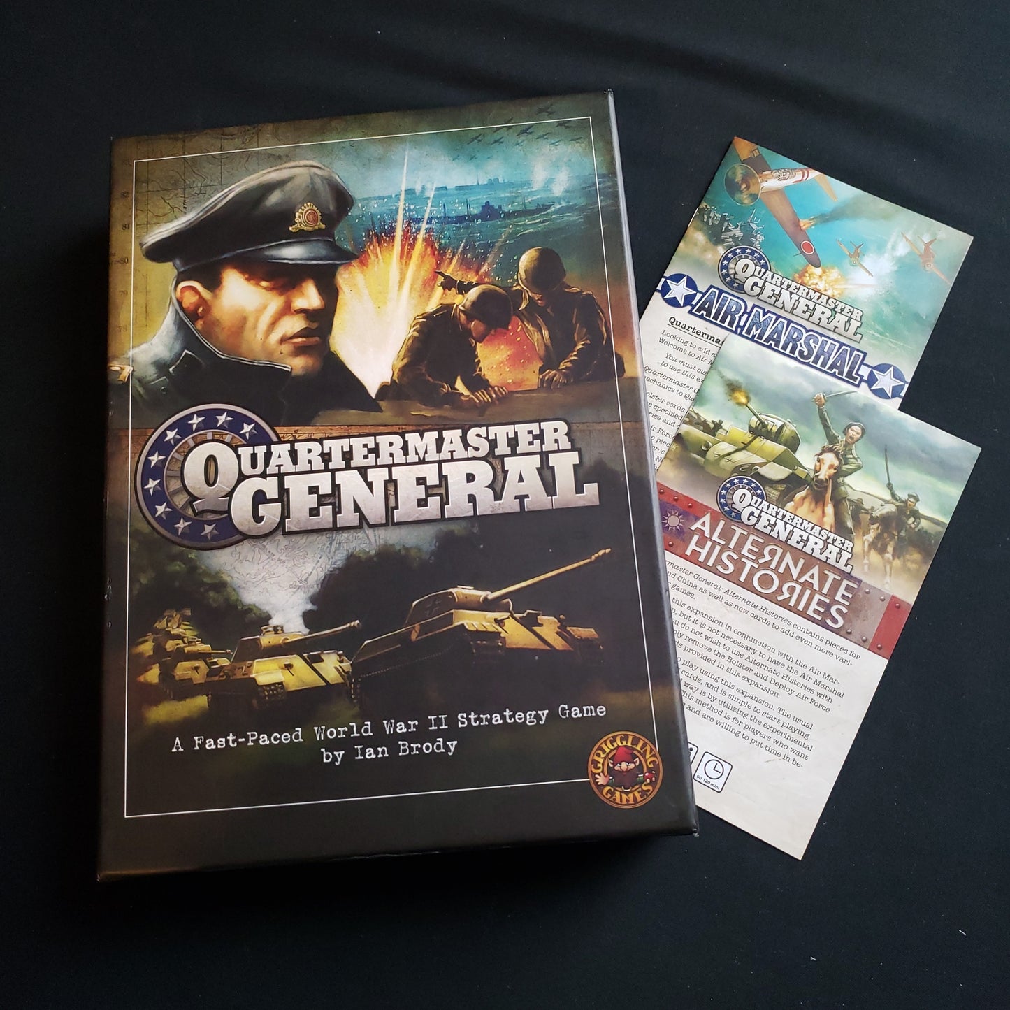 Image shows the front cover of the box of the Quartermaster General board game, with the instructions for the Air Marshal and Alternate Histories expansions tucked underneath the box