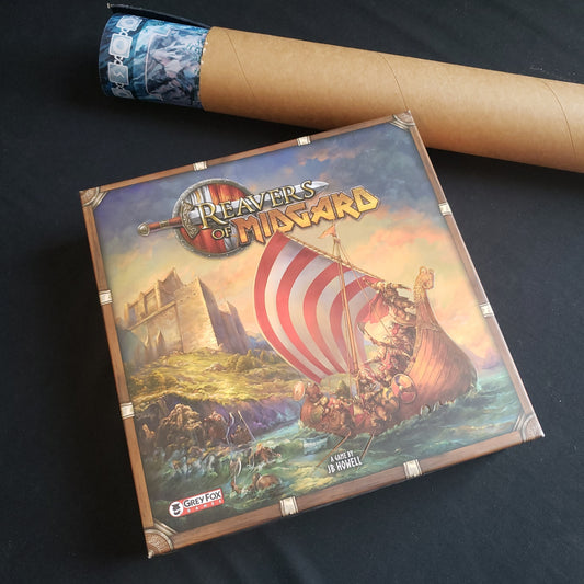Image shows the front cover of the box of the Reavers of Midgard board game, with the rolled up playmat in a tube above it