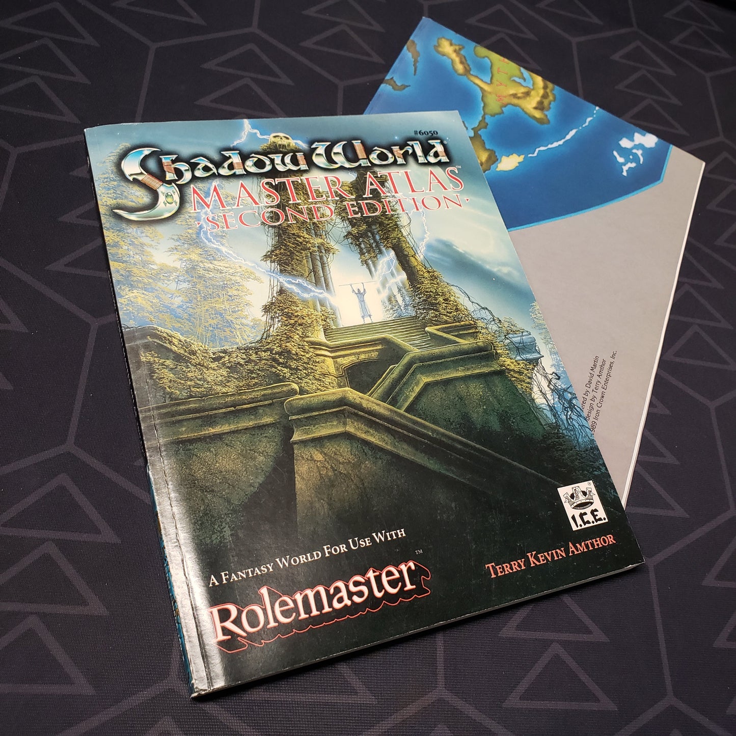Image shows the front cover of the Shadow World: Master Atlas - Second Edition book for the roleplaying game Rolemaster with the poster map sitting underneath it