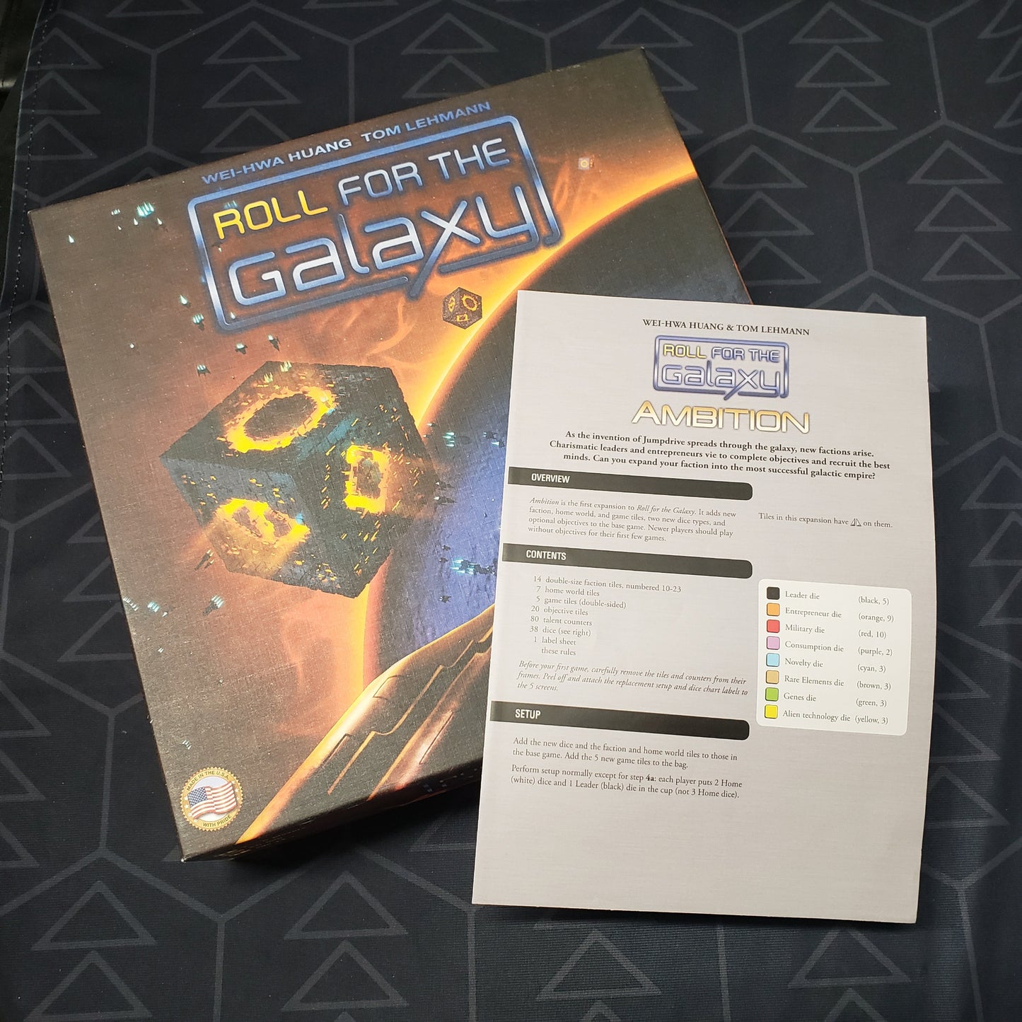 Image shows the front cover of the box of the Roll for the Galaxy board game with the instructions for the Ambition expansion sitting on top of it