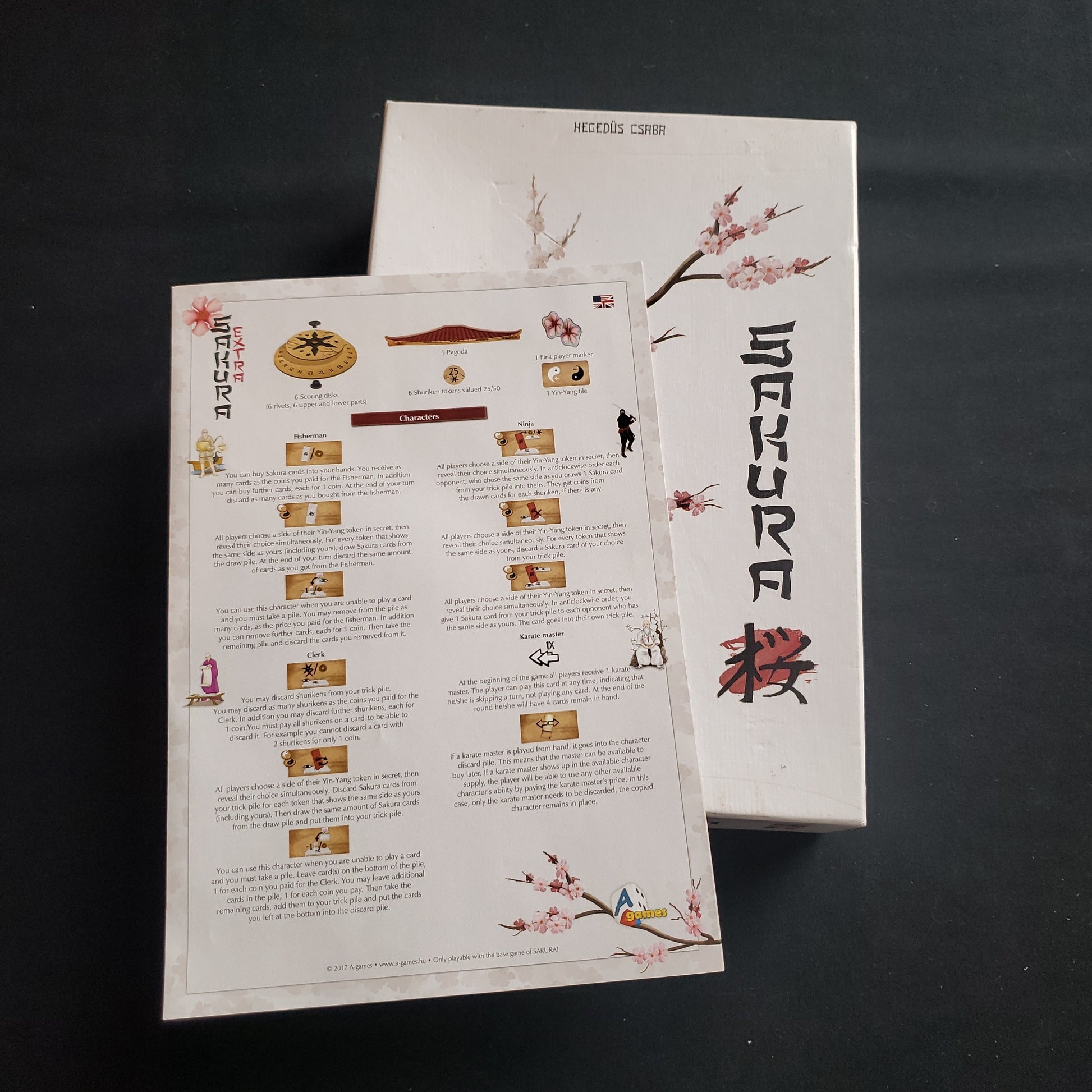 Image shows the front cover of the box of the Sakura board game, with the instructions for the Extras add-on on top of it