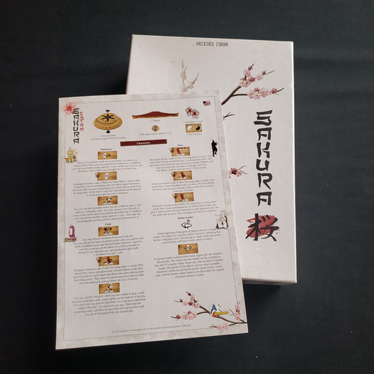 Image shows the front cover of the box of the Sakura board game, with the instructions for the Extras add-on on top of it