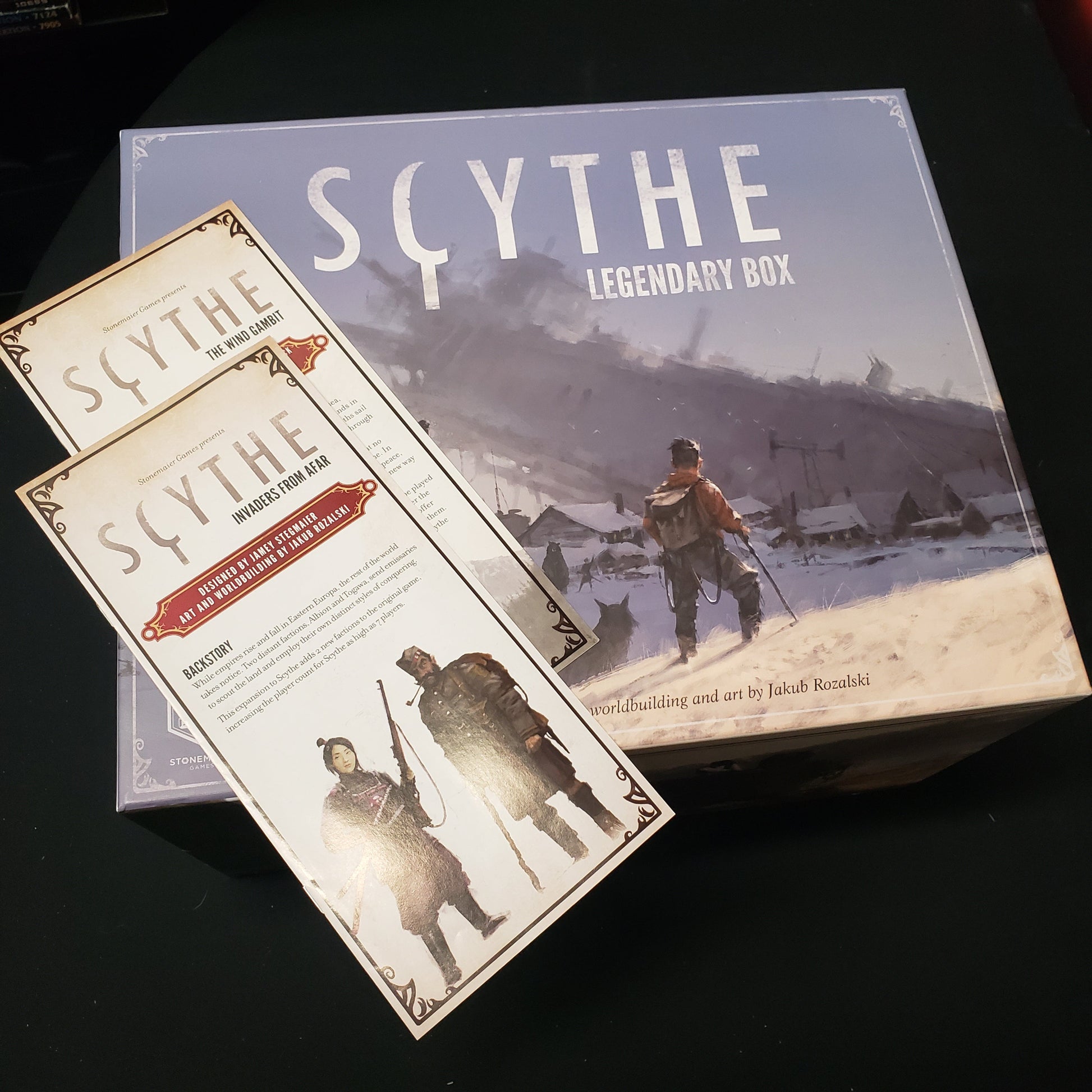 Image shows the front cover of the Legendary Box storage solution for the board game Scythe, with instructions for two expansions sitting on top of it