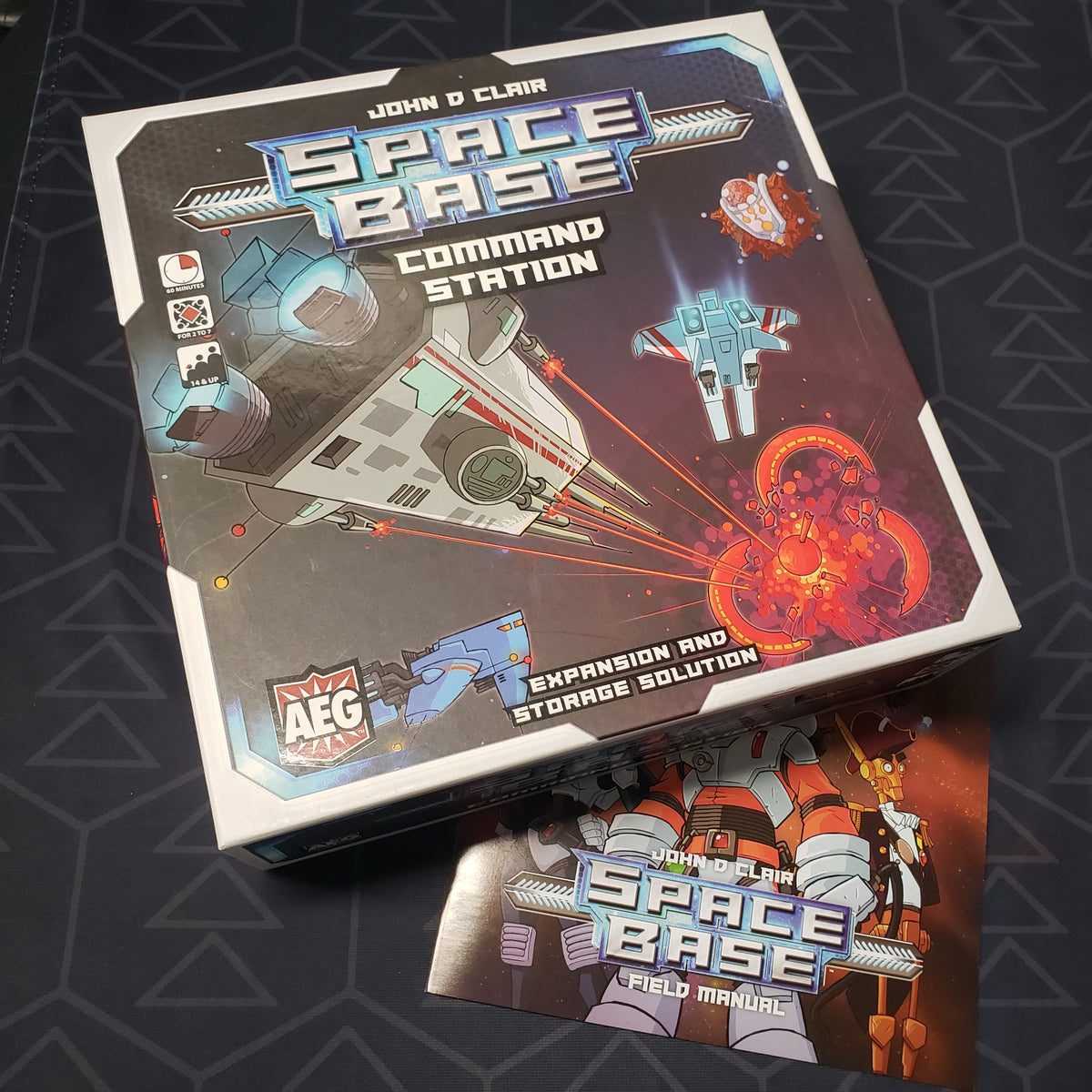 Image shows the front cover of the box of the Command Station expansion for the board game Space Base with the base game instructions sitting next to it