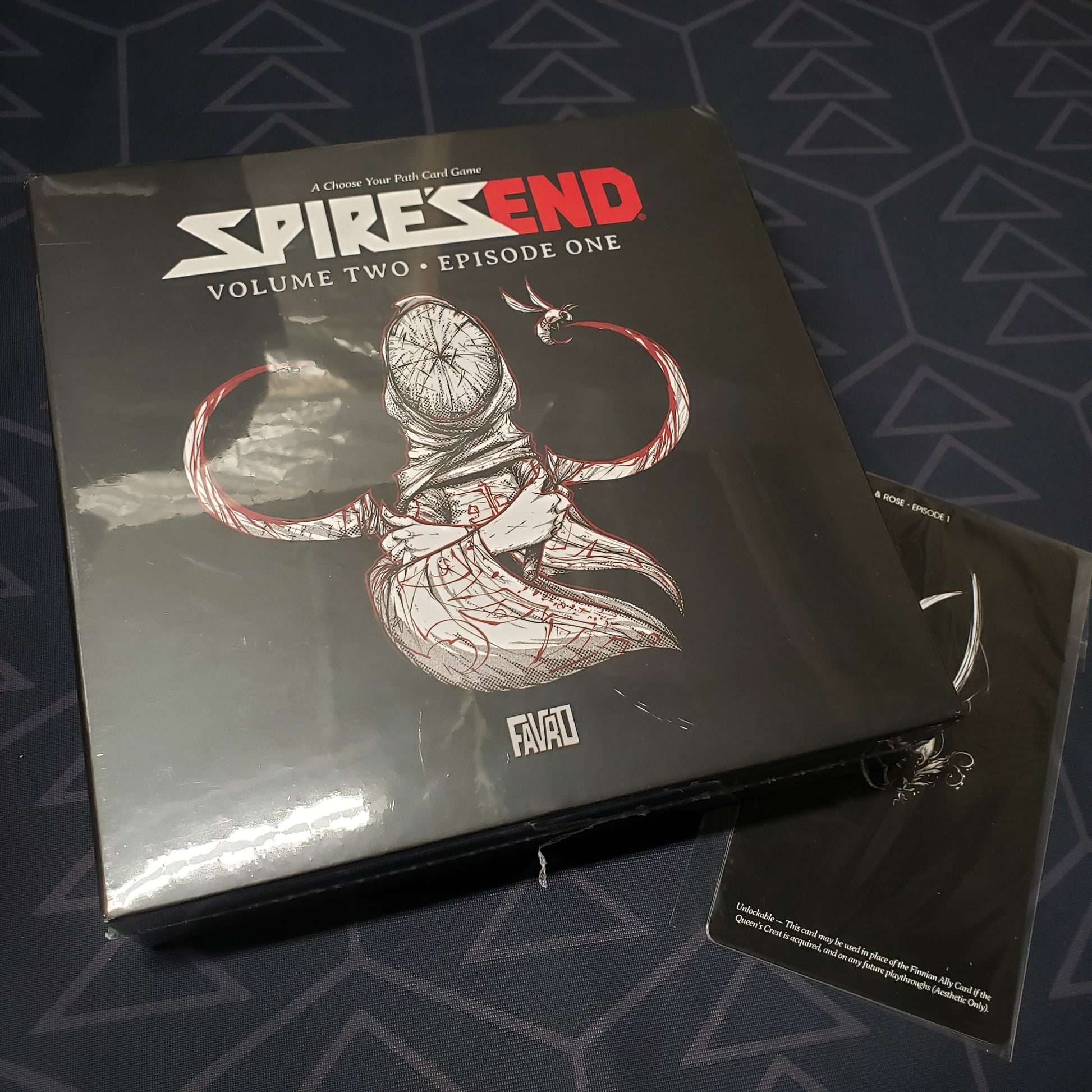 Image shows the front cover of the box for the Spire's End: Volume 2, Episode 1 board game sitting on top of a promo card pack