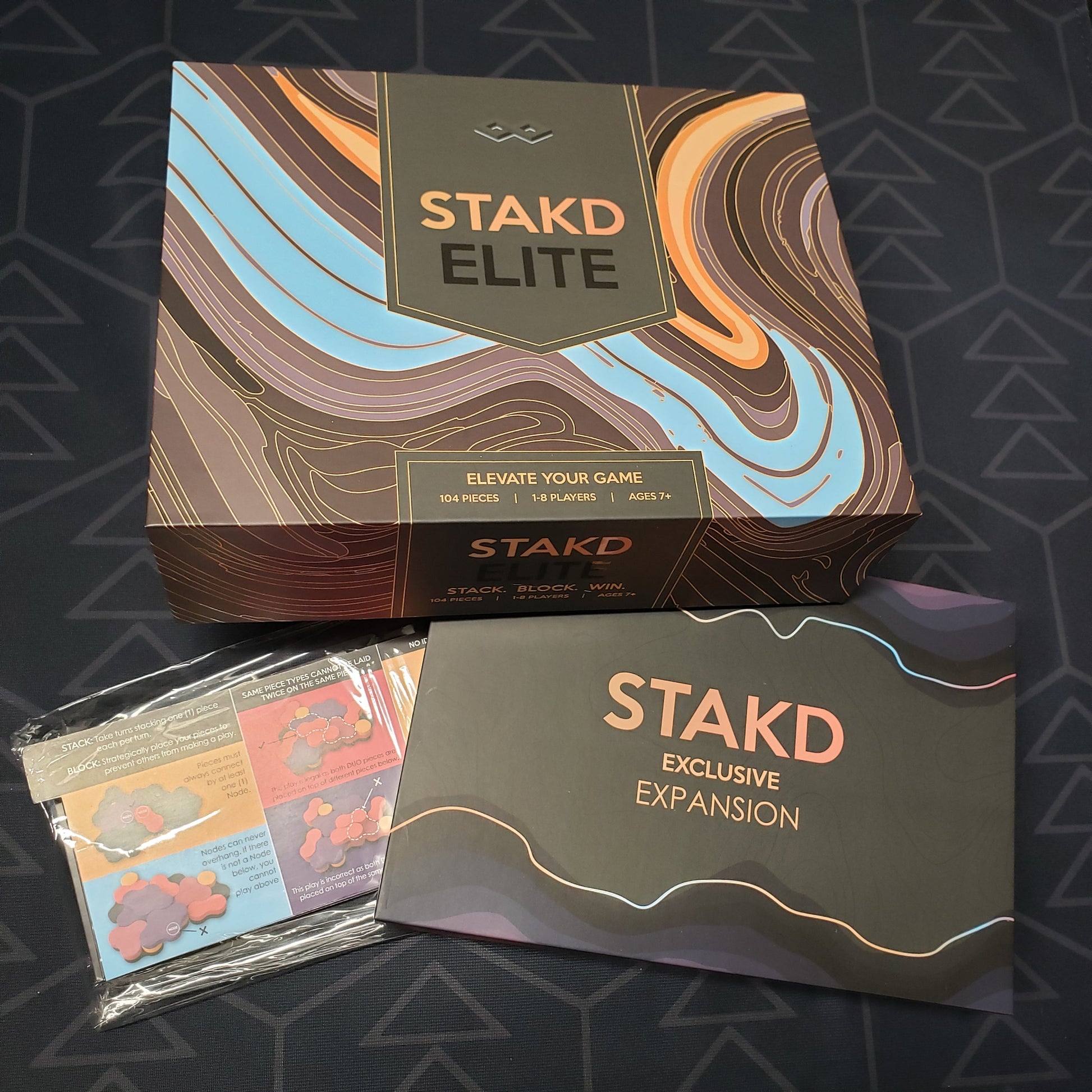 Image shows the front cover of the box for the STAKD Elite board game, along with the box for the Exclusive Expansion