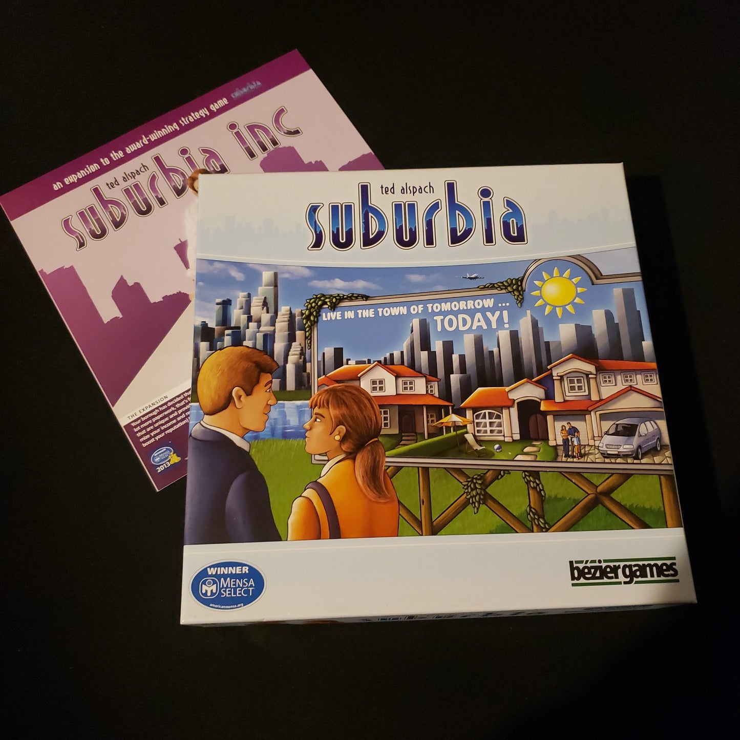 Image shows the front cover of the box of the Suburbia board game, sitting onto the instructions for the Suburbia Inc expansion