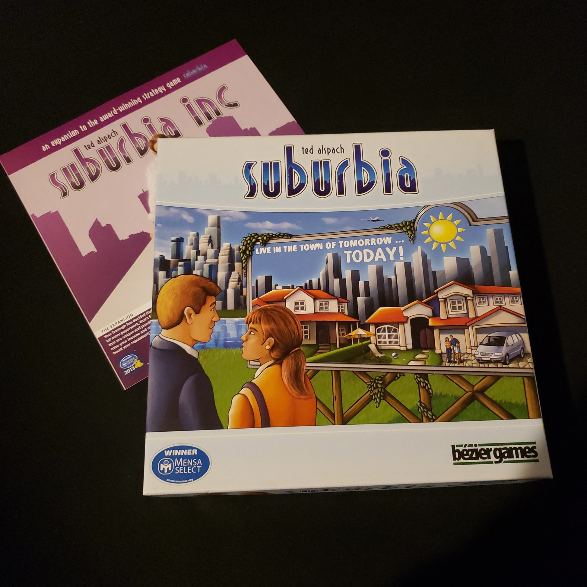 Image shows the front cover of the box of the Suburbia board game, sitting onto the instructions for the Suburbia Inc expansion