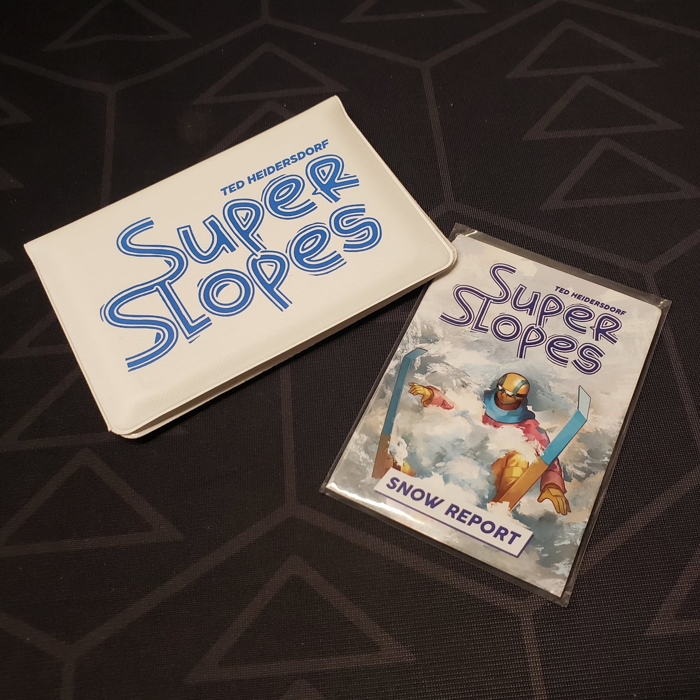 Image shows the front of the card wallet for the Super Slopes card game, with the Snow Report expansion sitting next to it