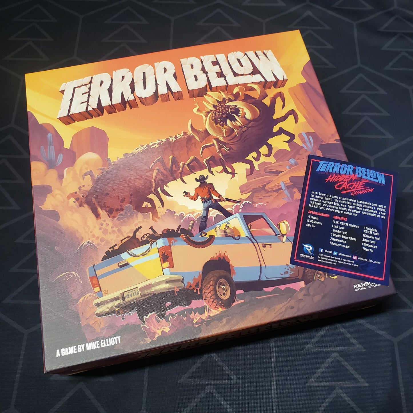 Image shows the front cover of the box for the Terror Below board game with the instructions for the Hidden Cache expansion sitting on top of it