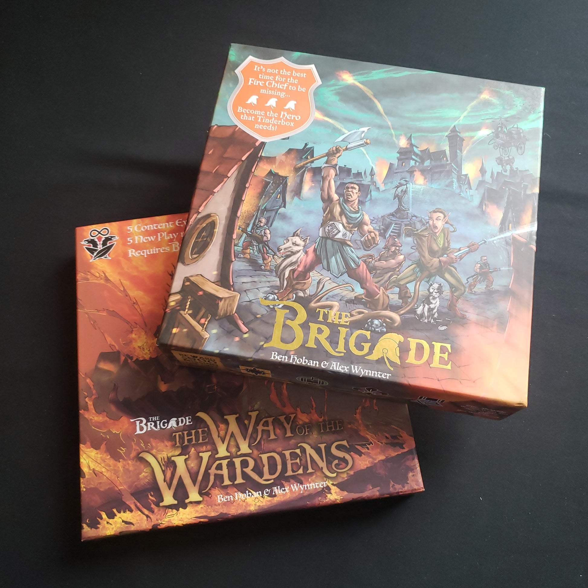 Image shows the box for the board game The Brigade sitting on top of the box for the Way of the Wardens expansion