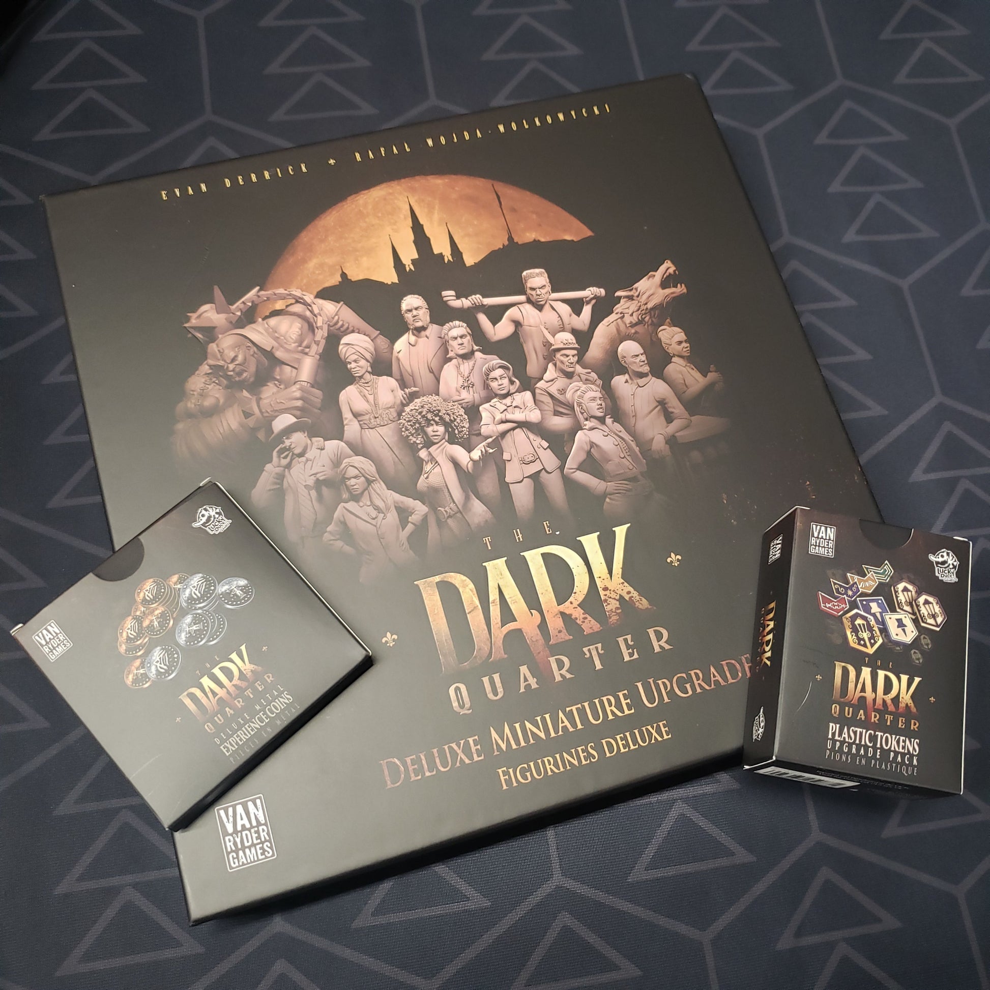 Image shows the front cover of the box of the Deluxe Miniature Upgrade expansion for the board game The Dark Quarter, with two smaller component boxes sitting on top of it