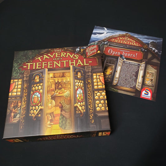 Image shows the front cover of the box of the Taverns of Tiefenthal board game, sitting on top of the instructions for the Open Doors expansion