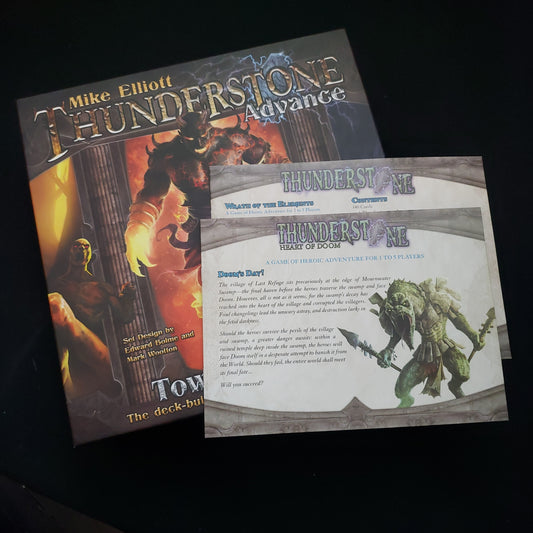 Image shows the front cover of the box of the Thunderstone Advance: Towers of Ruin board game, with the instructions for the original base game and Heart of Doom expansion sitting on top of it