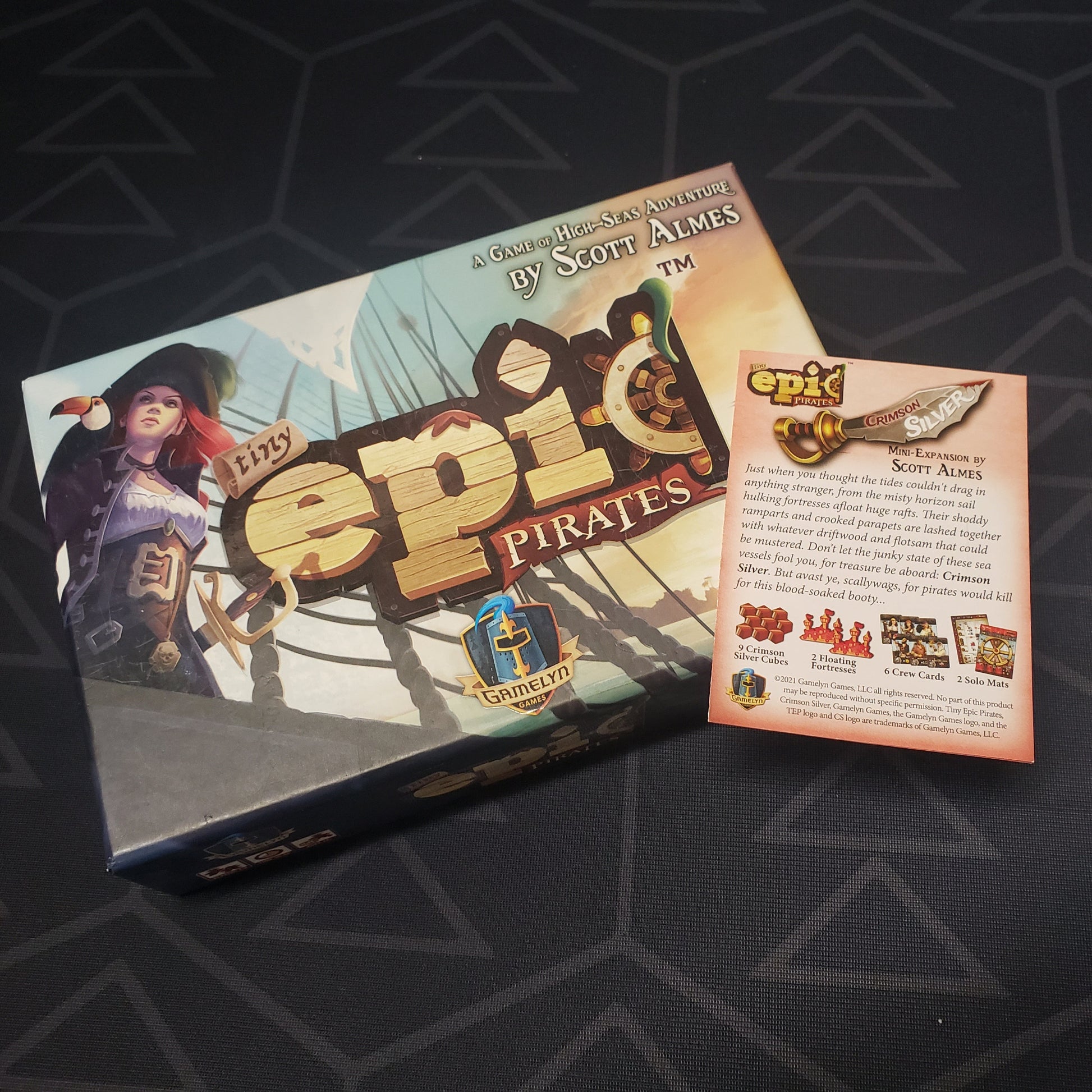 Image shows the front cover of the box of the Tiny Epic Pirates board game with the instructions for the Crimson Silver mini expansion sitting on top of it