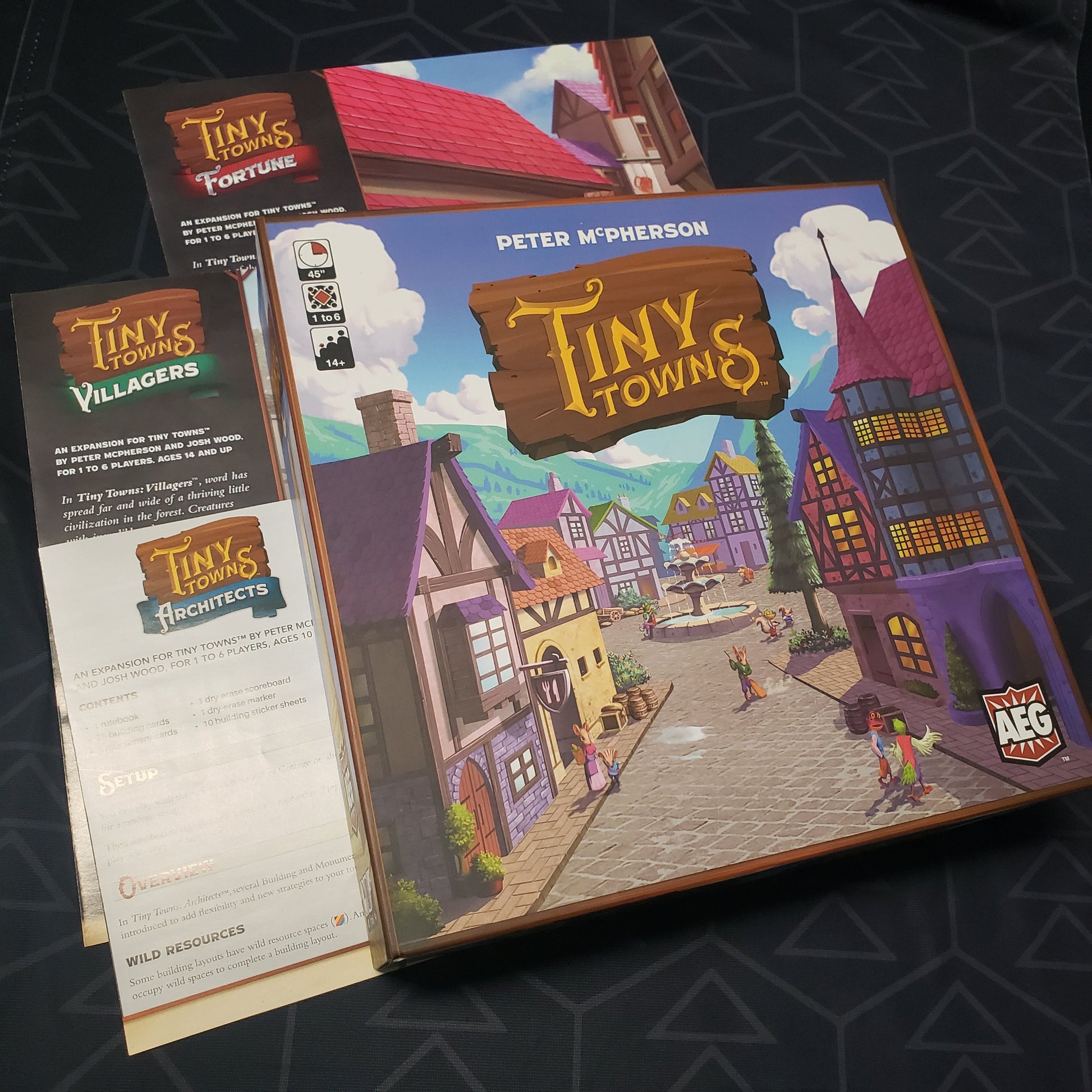 Image shows the front cover of the box of the Tiny Towns board game with three expansion manual sitting around it