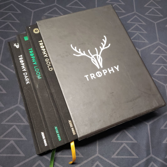 Image shows the front of the box for the Trophy: Hardcover Collection roleplaying game set, with the Dark, Gold & Loom books fanned out from the box