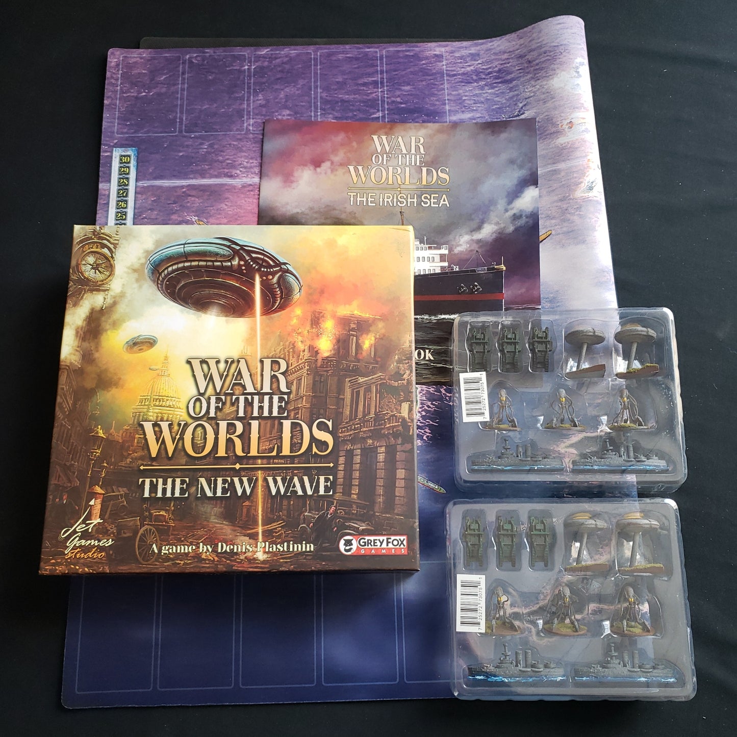 Image shows the box for the War of the Worlds: The New Wave board game sitting on top of the game's playmat. The instructions for the Irish Sea expansion and 2 sets of miniatures are sitting next to it