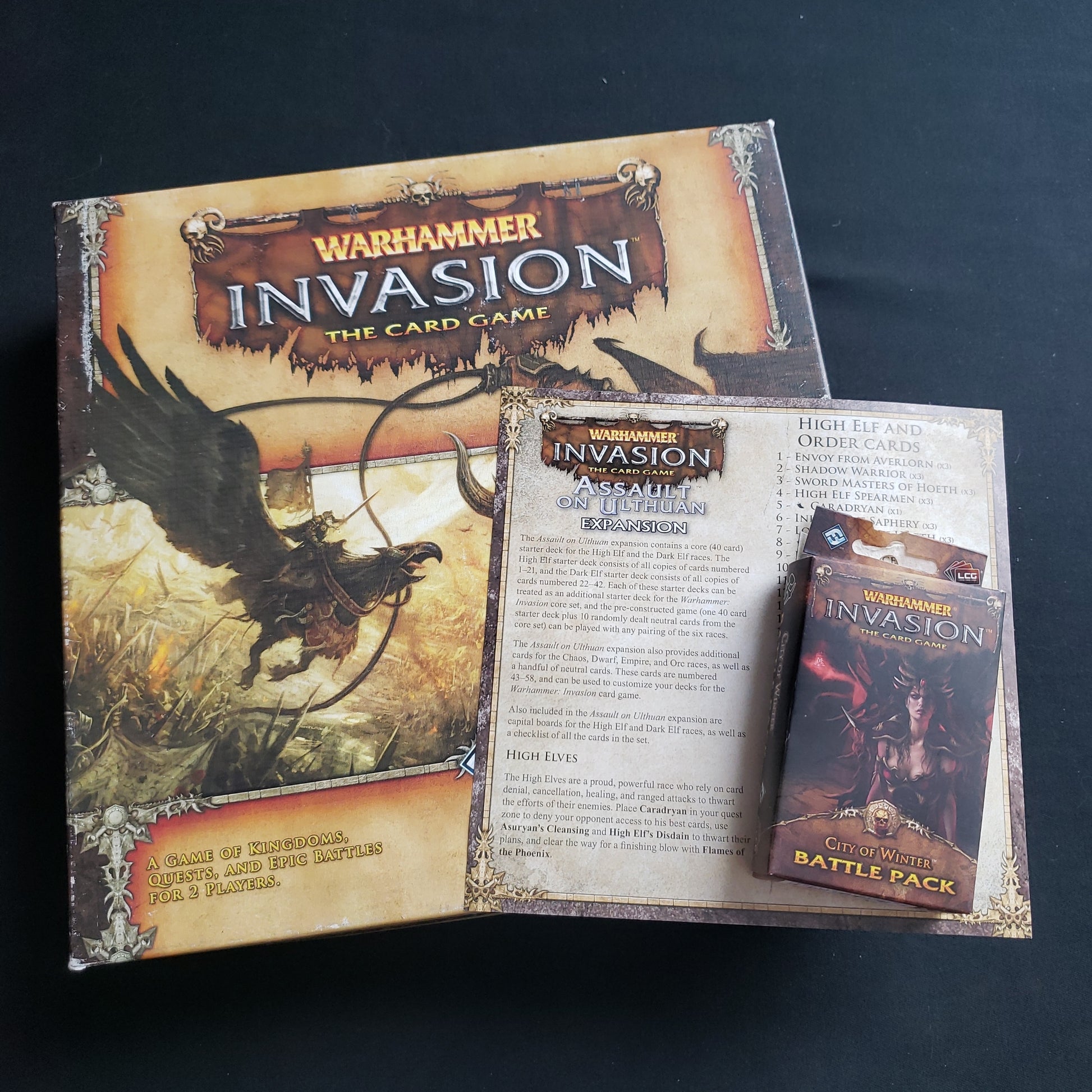 Image shows the front cover of the box of the Warhammer Invasion card game, with the instructions for the Assault on Ulthuan expansion and the packaging for the City of Winter Battle Pack sititng on top of it