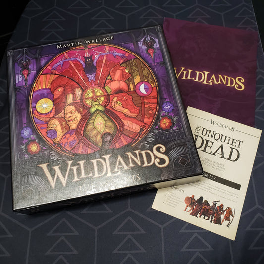 Image shows the front cover of the box of the Ancients expansion for the board game Wildlands sitting on top of the instructions for the base game and the Unquiet Dead expansion