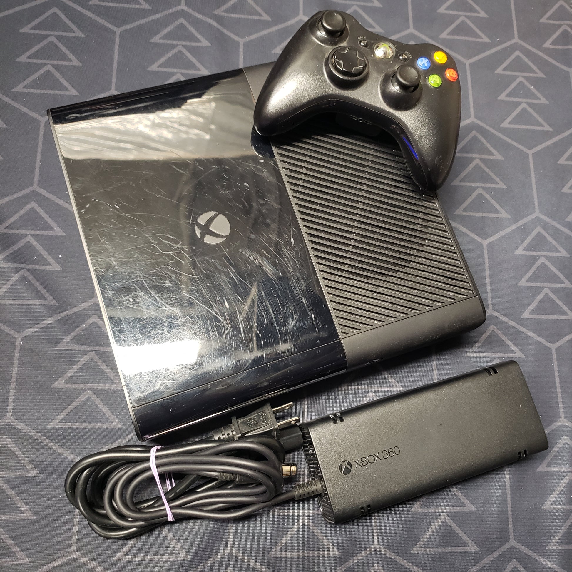 Image shows the top of a Xbox 360 E gaming console with a controller sitting on top and power supply sitting beside it