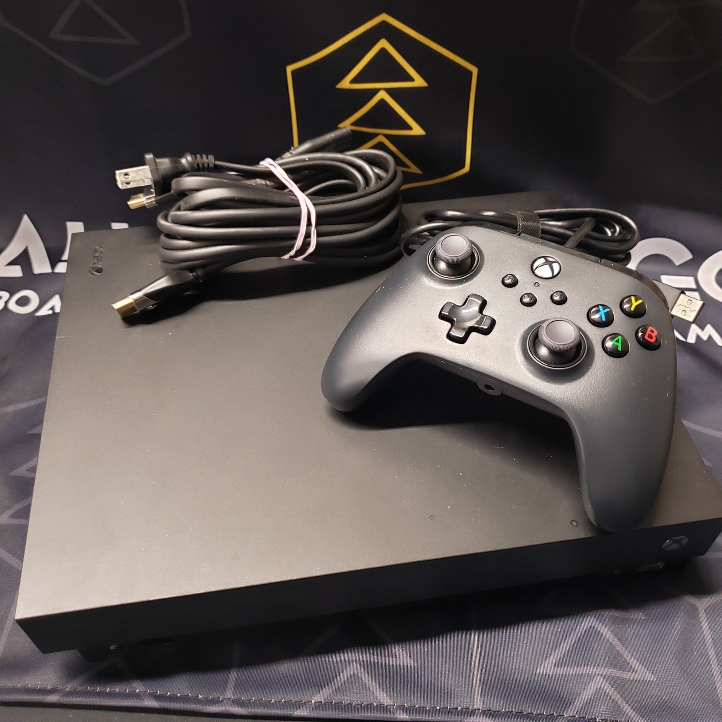 Image shows the top of a Xbox One X gaming console with a wired controller and cables wrapped up on top of it