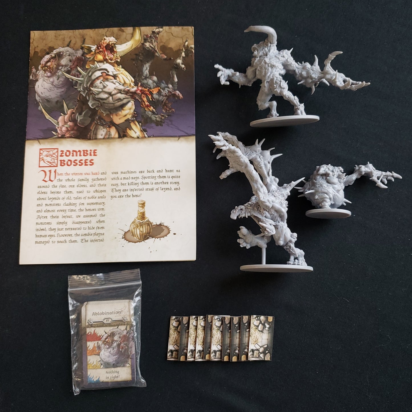 Image shows the instructions, miniatures and cards for the Zombie Bosses Abomination Pack for the board game Zombicide: Black Plague