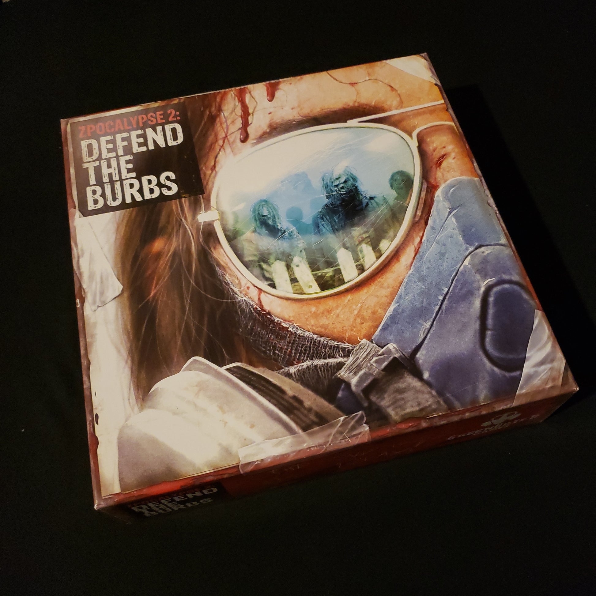 Image shows the front cover of the box of the Zpocalypse 2: Defend The Burbs board game