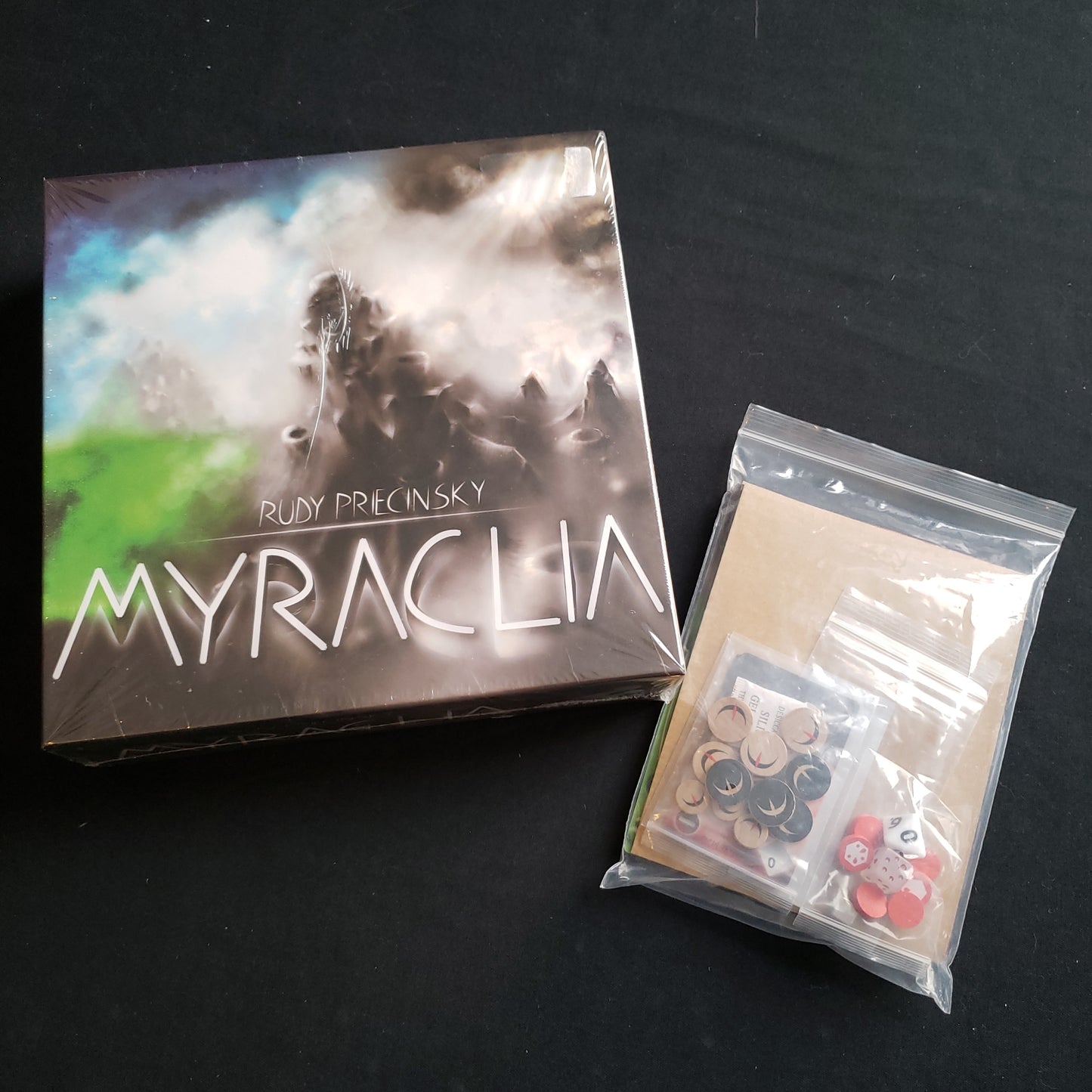 Image shows the front cover of the box of the Myraclia board game, along with the Luna expansion in a clear plastic bag next to it